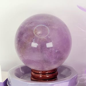 May include: A large, polished amethyst crystal ball, resting on a small, dark wooden stand. The sphere is a translucent purple color with internal patterns. It sits atop a lavender container, with a small box that says "Play" in the foreground.