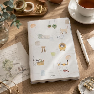 May include: A 2027 datebook planner with a white cover featuring illustrations of cats in various poses, including sleeping, walking, and sitting in a basket. The cover includes thin line drawings of furniture and plants, with the text 2027 Datebook.