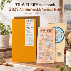 May include: A mustard yellow Traveler's notebook 2027 A5 slim weekly vertical refill with a white label featuring the text 2027 Weekly Vertical. Beside it is a light peach 2027 guide booklet with various small illustrations and a tan sticker sheet.