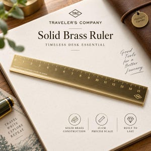 Op de afbeelding: Een liniaal van massief messing met een schaalverdeling van 15 cm, voorzien van de tekst Traveler's Company Made in Japan. De liniaal ligt op een pagina met de tekst Traveler's Company Solid Brass Ruler Timeless Desk Essential en Good Tools for a Better Journey.
