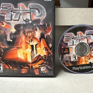 Pode incluir: Caixa e disco do jogo "Robot Alchemic Drive" para PlayStation 2. A caixa apresenta uma ilustração de um robô com um fundo de cidade. O disco mostra o título do jogo e o logotipo do PlayStation 2.