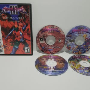 Pode incluir: Um conjunto de videojogos Sega Saturn com "Shining Force III: Scenario 1, 2 & 3". O conjunto inclui a caixa do jogo e quatro discos de jogo. A caixa tem uma capa vermelha e azul com o título do jogo e o texto "SEGA SATURN".