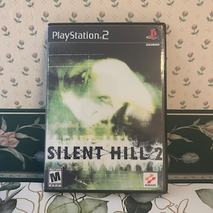 Puede incluir: Caja de videojuego de PlayStation 2 para Silent Hill 2. La portada presenta una imagen borrosa y fantasmal en tonos verdes y blancos. El título del juego se muestra en letras grandes y en negrita. La caja está sellada en plástico.
