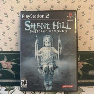 May include: A PlayStation 2 video game case for "Silent Hill: Shattered Memories." The cover art features a ghostly figure on a swing set, with the game's title in a stylized font. The case is black with the PlayStation 2 logo.