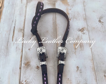 May include: Brown leather horse headstall with a pink diamond stitch pattern and silver floral conchos.  "Lucky Leather Company" is printed on the background.