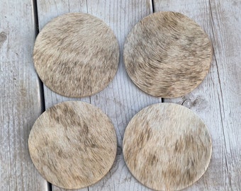 May include: Set of four brown and white cowhide coasters. The coasters are round and have a natural, rustic look.  The coasters are arranged in a square pattern on a wooden surface.  The text "Lucky Leather Company" is visible in the bottom center of the image.
