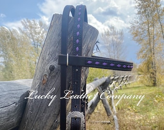 May include: A brown leather horse halter with purple diamond stitching and a silver buckle. The halter is hanging on a wooden fence post. "Lucky Leather Company" is written on the post.
