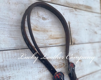 May include: A black leather horse headstall with red and copper accents. The headstall has a diamond pattern and two decorative buckles.