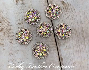 May include: Six silver-toned round conchos with iridescent pink and clear rhinestones. The conchos are arranged in a semi-circle on a light wood surface. The text "Lucky Leather Company" is visible in the bottom right corner of the image.
