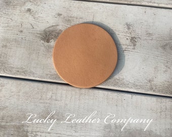 May include: A round, light brown leather coaster. The coaster is on a white wooden surface. The text "Lucky Leather Company" is written in white cursive below the coaster.
