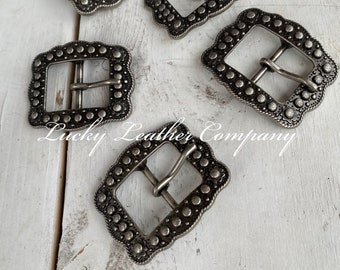 Op de afbeelding: Vijf zilverkleurige metalen gespen met een decoratief ontwerp. Elke gesp heeft een vierkante vorm met afgeronde hoeken en een verhoogde rand. De gespen hebben een getextureerd oppervlak met kleine, verhoogde stippen.