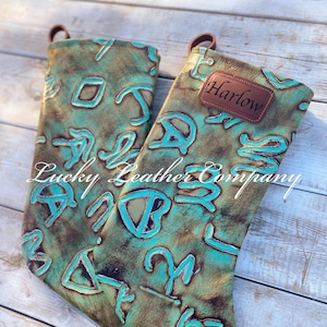 May include: Two turquoise and brown leather stockings with a western design. The stocking on the right has a brown leather tag with the name "Harlow" on it. The stockings are from Lucky Leather Company.