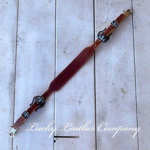 May include: A brown leather strap with two decorative metal buckles. The buckles have a blue and silver design. The strap is on a white wooden surface. "Lucky Leather Company" is written in white text on the surface.
