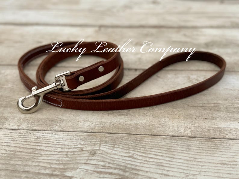 Peut inclure: Une laisse de chien en cuir marron avec une attache argent&eacute;e et des coutures. La laisse est sur une surface en bois. Lucky Leather Company est &eacute;crit en blanc sur la surface.