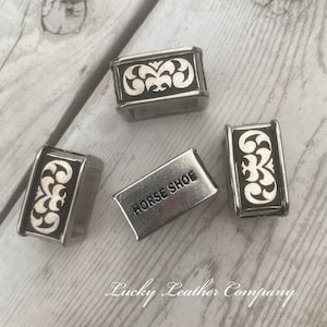 May include: Four silver metal belt buckle slides. Three have a decorative design with a black background and white swirls. One slide has the words "HORSE SHOE" engraved on it.