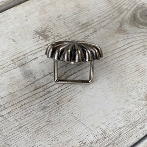 May include: Silver-toned metal belt buckle with a decorative, sunburst-like design. The buckle has a square frame.