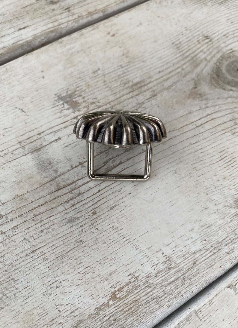 May include: Silver-toned metal belt buckle with a decorative, sunburst-like design. The buckle has a square frame.