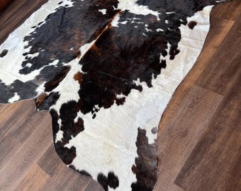 May include: A cowhide rug with a natural pattern of black and brown spots on a white background. The rug is irregularly shaped and lies on a dark wood floor. The rug's texture and color variations are visible.