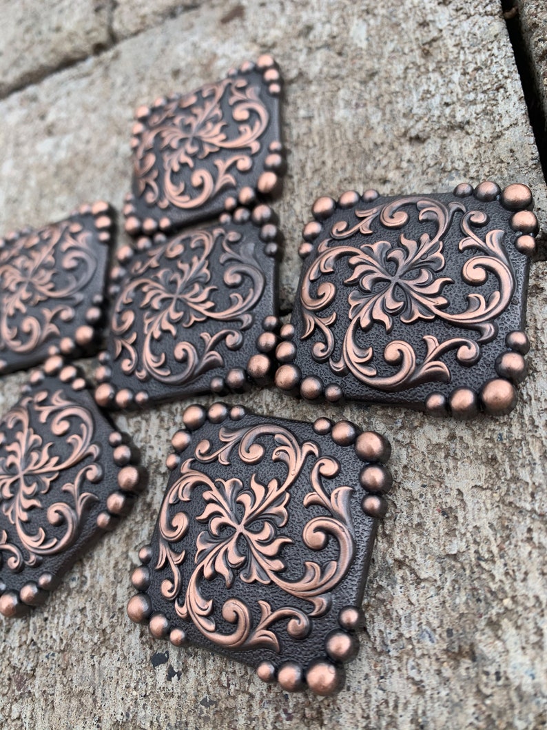Puede incluir: Primer plano de varios adornos met&aacute;licos cuadrados de color bronce. Cada uno tiene un dise&ntilde;o floral en relieve detallado en un tono bronce m&aacute;s claro. Los bordes est&aacute;n bordeados con peque&ntilde;os acentos redondos de bronce, creando una apariencia decorativa y ornamentada. Probablemente se trate de herrajes decorativos.