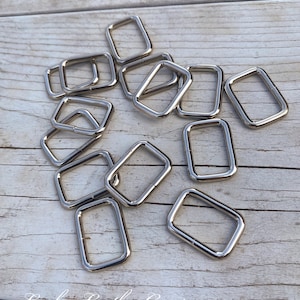 May include: A group of silver metal D-rings, scattered on a light wood surface.  The rings are all the same size and shape, and they are arranged in a random pattern.  The rings are a common hardware item used in leather crafting and other DIY projects.