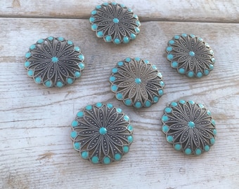 May include: Six turquoise and silver round buttons with a floral design. The buttons are arranged in a semi-circle on a light wood background.