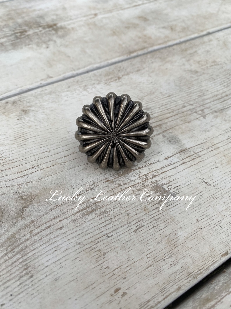 May include: A silver metal knob with a sunburst design. The knob is on a white wooden surface.