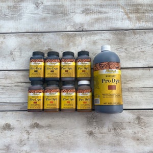 K&ouml;nnte beinhalten: Ein Set mit 10 Fiebings Pro Dye Lederfarbflaschen in verschiedenen Farben, darunter Schwarz, Schokolade, Dunkelbraun, Mahagoni, Englisch Zaumzeug, Hellbraun und Sattelbraun. Die Flaschen sind in zwei Reihen auf einer Holzoberfl&auml;che angeordnet. Die Flaschen haben jeweils einen Inhalt von 4 Fl&uuml;ssigunzen. Es gibt auch eine gr&ouml;&szlig;ere 32 Fl&uuml;ssigunzen Flasche Fiebings Mahogany Pro Dye.