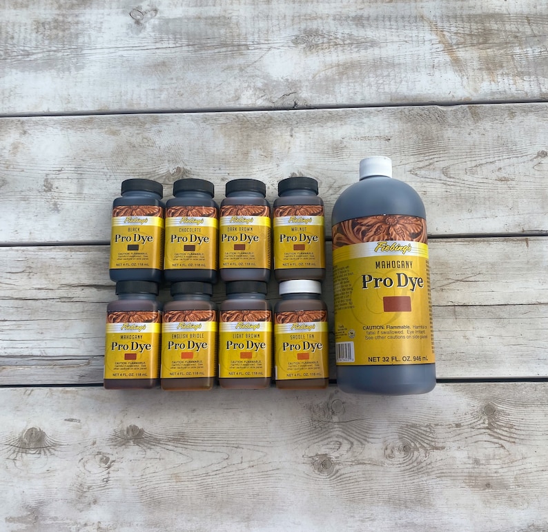 K&ouml;nnte beinhalten: Ein Set mit 10 Fiebings Pro Dye Lederfarbflaschen in verschiedenen Farben, darunter Schwarz, Schokolade, Dunkelbraun, Mahagoni, Englisch Zaumzeug, Hellbraun und Sattelbraun. Die Flaschen sind in zwei Reihen auf einer Holzoberfl&auml;che angeordnet. Die Flaschen haben jeweils einen Inhalt von 4 Fl&uuml;ssigunzen. Es gibt auch eine gr&ouml;&szlig;ere 32 Fl&uuml;ssigunzen Flasche Fiebings Mahogany Pro Dye.