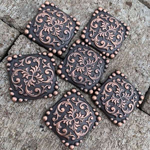 Puede incluir: Seis adornos cuadrados decorativos de metal color cobre con un dise&ntilde;o floral ornamentado. Cada cuadrado est&aacute; enmarcado con peque&ntilde;os tachuelas redondos de color cobre. Los adornos est&aacute;n dispuestos sobre una superficie gris texturizada.