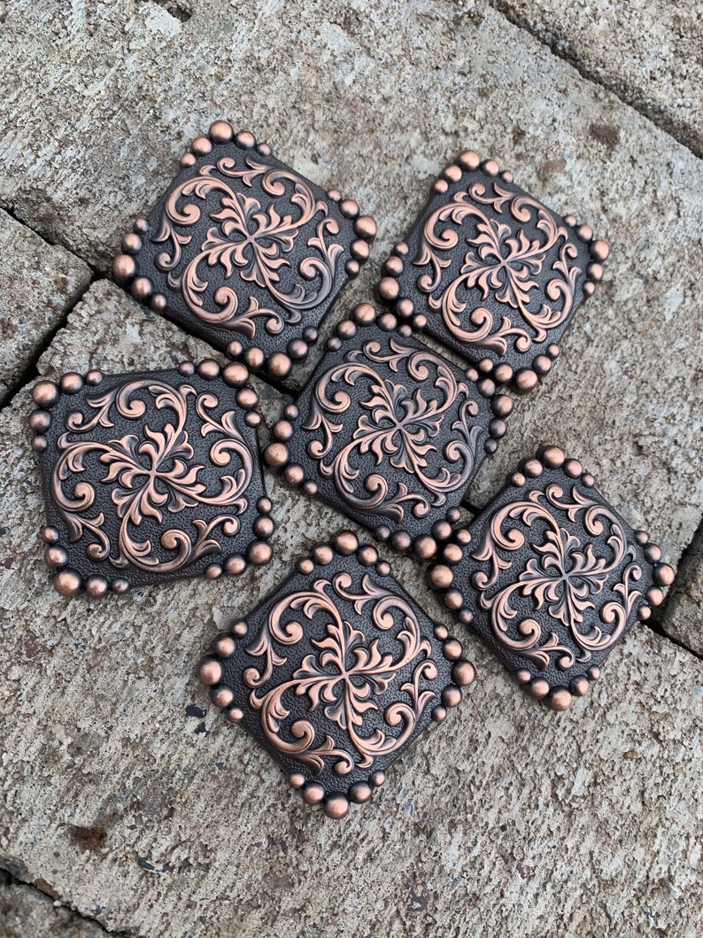Puede incluir: Seis adornos cuadrados decorativos de metal color cobre con un dise&ntilde;o floral ornamentado. Cada cuadrado est&aacute; enmarcado con peque&ntilde;os tachuelas redondos de color cobre. Los adornos est&aacute;n dispuestos sobre una superficie gris texturizada.
