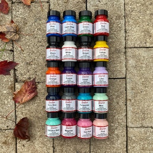 Puede incluir: Una colecci&oacute;n de 18 peque&ntilde;os frascos de pintura acr&iacute;lica para cuero Angelus en varios colores, incluyendo azul, verde, rojo, negro, blanco, marr&oacute;n, amarillo, naranja, morado, violeta, lila, azul marino, gris, turquesa, menta, azul caja de regalo, magenta, rosa intenso y rosa. Los frascos est&aacute;n dispuestos en cuatro filas de cuatro frascos cada una, y todos est&aacute;n sobre una superficie de piedra.