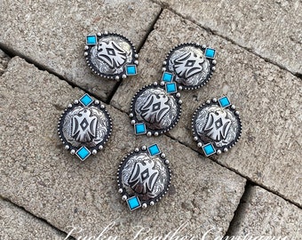 May include: Six silver conchos with turquoise stones. Each concho features a stylized design with a diamond shape in the center. The conchos are arranged in a circle on a gray brick surface.