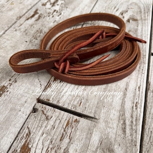 May include: Coiled brown leather reins with a looped end and knotted details. The reins are made of a rich, reddish-brown leather and appear to be handmade. The image is set against a rustic, weathered wooden background.