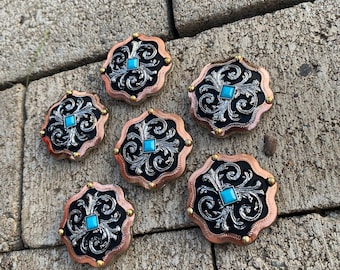 May include: Six decorative metal buttons with a black background, silver filigree, and a turquoise stone in the center. The buttons are arranged in a circle on a gray stone surface.