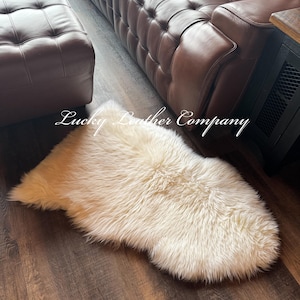 K&ouml;nnte beinhalten: Cremefarbener Schaffellteppich auf einem Holzboden. Der Teppich hat eine weiche, flauschige Textur und befindet sich in der N&auml;he eines braunen Ledersofas und einer Ottomane. Der Text "Lucky Leather Company" ist sichtbar.
