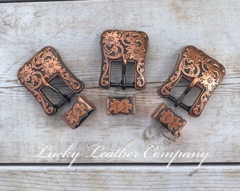 May include: Three antique copper-toned belt buckles with intricate floral designs. Each buckle has a black metal tongue and is accompanied by a matching copper-toned slider. The buckles are arranged on a light wood surface with the text "Lucky Leather Company" visible below.