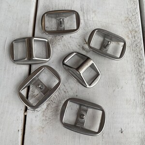 May include: Six silver metal belt buckles with a rectangular shape and a small hole in the center.