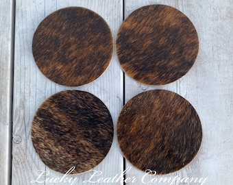 May include: Four brown and black cowhide coasters with a light brown background. The coasters are arranged in a square pattern on a white wooden surface. The text "Lucky Leather Company" is visible in the bottom center of the image.