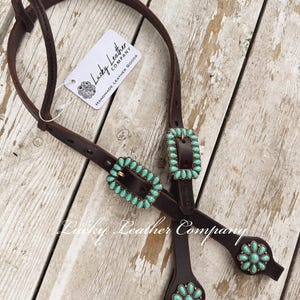 May include: A dark brown leather horse headstall featuring two rectangular buckles and two circular conchos, each inlaid with multiple teardrop-shaped turquoise stones. A white tag attached to the leather reads Lucky Leather Company.