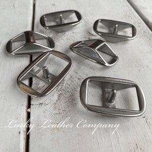 May include: Six silver metal belt buckle loops, also known as strap keepers, on a white wooden surface.  The loops are rectangular with a rounded edge and a small metal pin in the center.