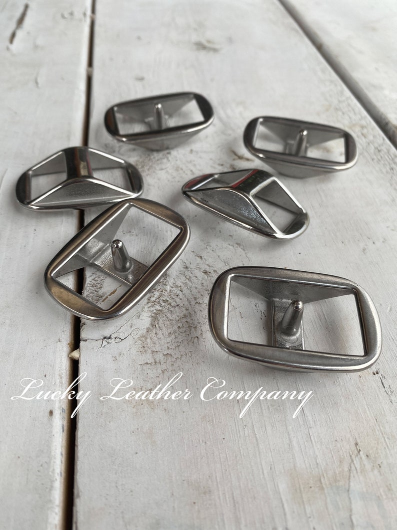 May include: Six silver metal belt buckle loops, also known as strap keepers, on a white wooden surface.  The loops are rectangular with a rounded edge and a small metal pin in the center.