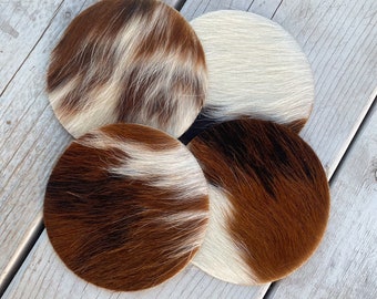 May include: Four round cowhide coasters in brown, white, and black. The coasters are arranged in a circle on a wooden surface. The text "Lucky Leather Company" is visible in the bottom of the image.