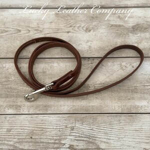 Peut inclure: Une laisse de chien en cuir marron avec un fermoir argent&eacute;. La laisse est enroul&eacute;e sur une surface en bois. Le texte "Lucky Leather Company" est visible en arri&egrave;re-plan.