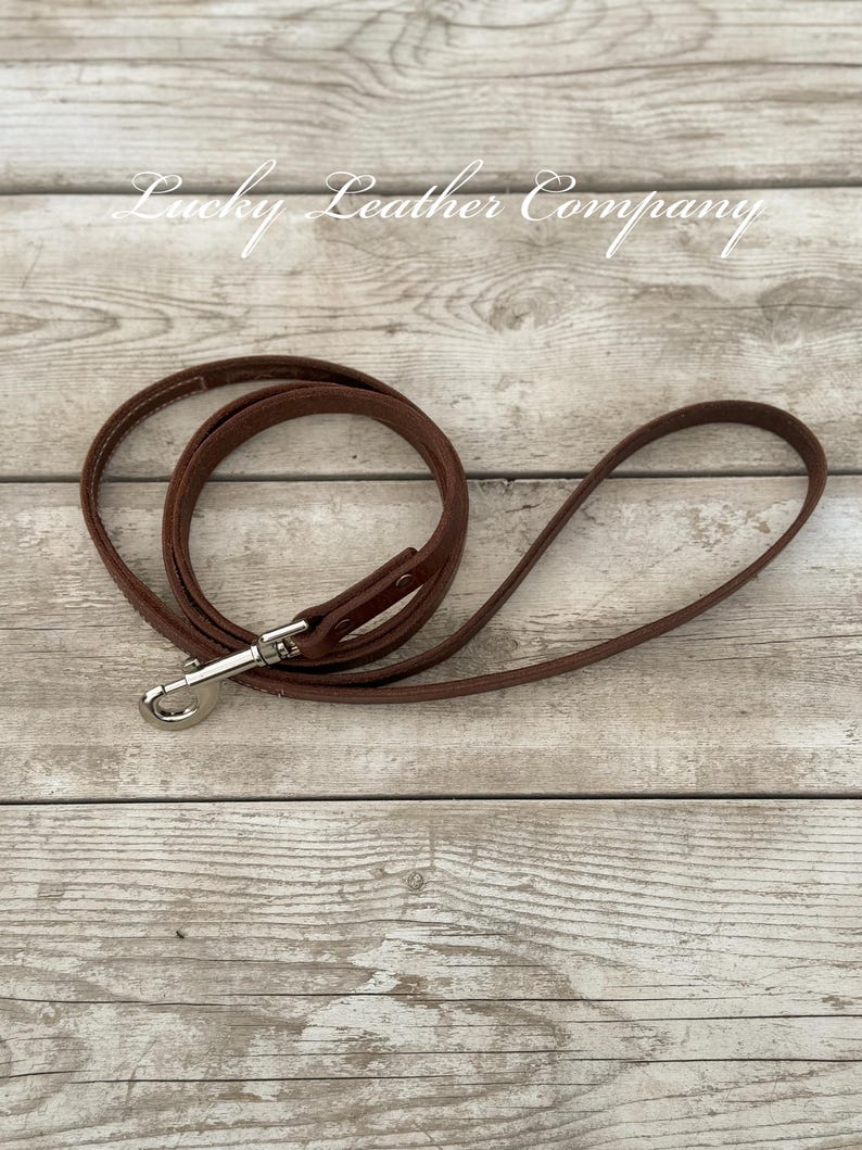 Peut inclure: Une laisse de chien en cuir marron avec un fermoir argent&eacute;. La laisse est enroul&eacute;e sur une surface en bois. Le texte "Lucky Leather Company" est visible en arri&egrave;re-plan.