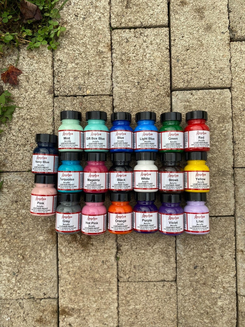 Puede incluir: Una colecci&oacute;n de 18 peque&ntilde;os frascos de pintura acr&iacute;lica para cuero Angelus en varios colores, incluyendo azul marino, turquesa, magenta, negro, blanco, marr&oacute;n, amarillo, rosa, gris, rosa intenso, naranja, morado, violeta y lila. Cada frasco tiene una etiqueta con el nombre del color y "Angelus Acrylic Leather Paint" impreso en ella.