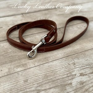 Peut inclure: Une laisse de chien en cuir marron avec un mousqueton argent&eacute;. La laisse est pos&eacute;e sur une surface en bois. Le texte "Lucky Leather Company" est visible en haut au centre de l'image.