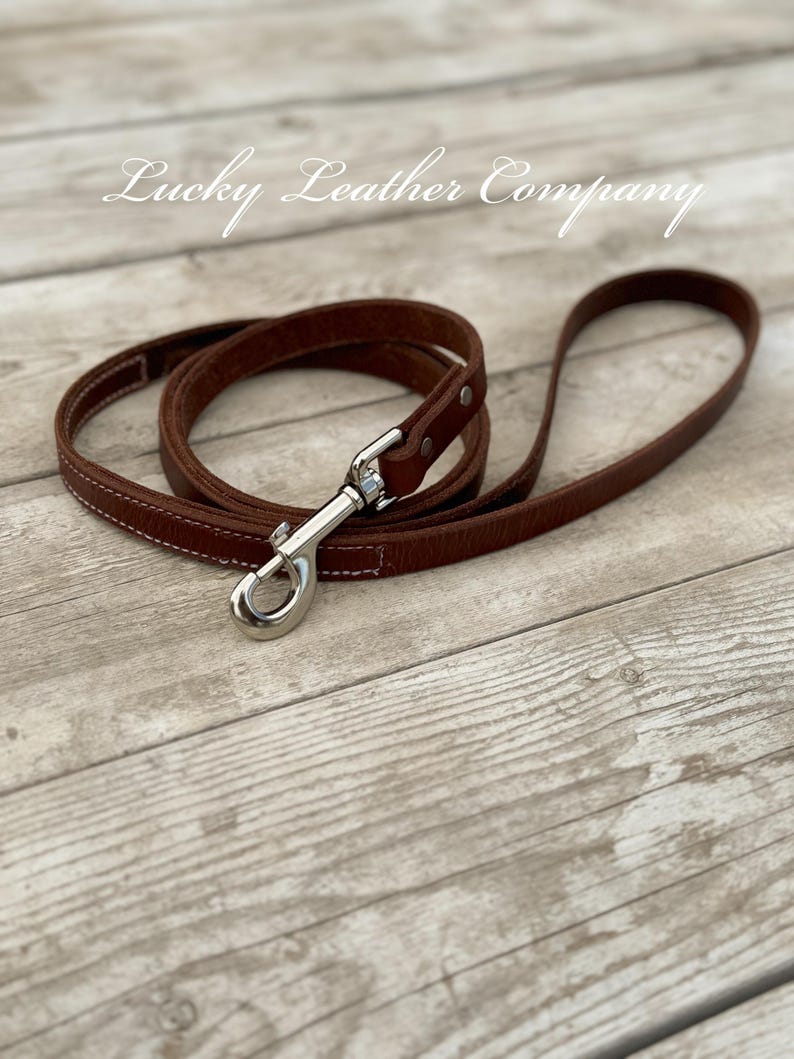 Peut inclure: Une laisse de chien en cuir marron avec un mousqueton argent&eacute;. La laisse est pos&eacute;e sur une surface en bois. Le texte "Lucky Leather Company" est visible en haut au centre de l'image.
