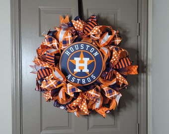 May include: A wreath decorated with orange, white, and blue ribbons featuring the Houston Astros logo. The logo is a blue circle with a white star and the text "HOUSTON ASTROS" in white. The letter "H" is in the center of the star.