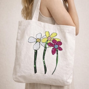 以下が含まれることがあります： 白いキャンバスのトートバッグ。3つの刺繍された花が特徴です。花は白、黄色、マゼンタの花びらで、スパンコールのディテールがあります。茎は緑色で、スパンコールのアクセントが付いています。バッグには白いハンドルが2つ付いています。