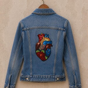 May include: A light blue denim jacket with a colorful sequined anatomical heart design on the back. The heart features red, blue, yellow, and green accents. The jacket is hanging on a wooden hanger.